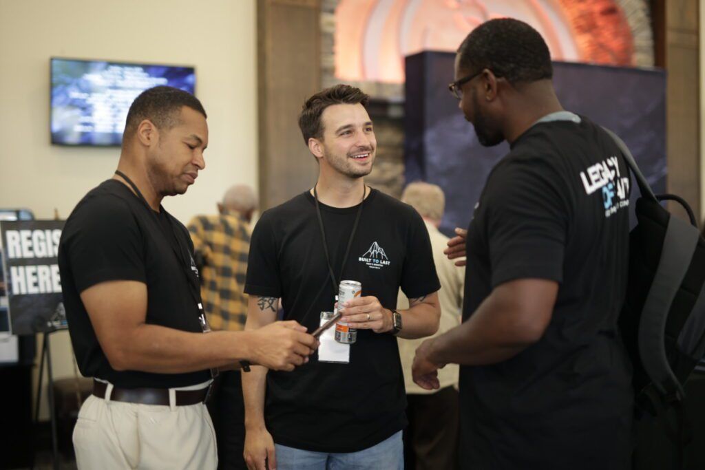 Three men chatting and smiling at an indoor event booth, holding drinks and lanyards