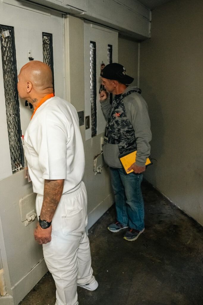Two people stand in a narrow hallway by barred doors, one holding a yellow folder.