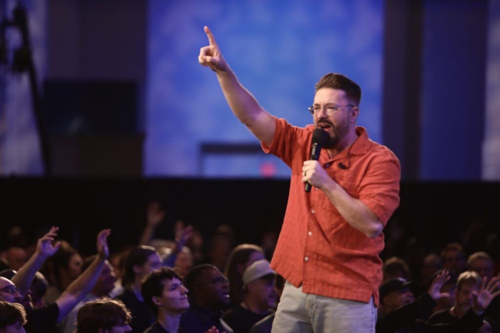 Singer in orange shirt raises one finger while performing to a cheering crowd under purple lights