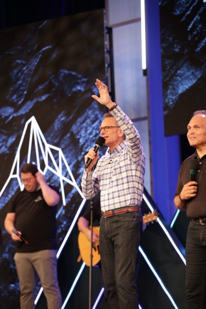 Three men onstage at an event, one speaking into a microphone with a raised hand.