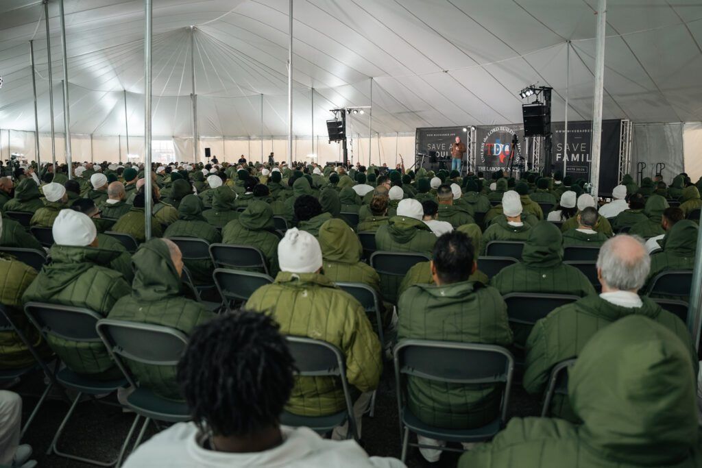 Audience seated in green chairs inside a white event tent, facing a stage with screens and lights