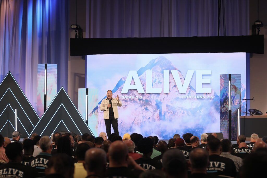 Speaker on stage in front of large “ALIVE” screen, with audience seated in foreground.