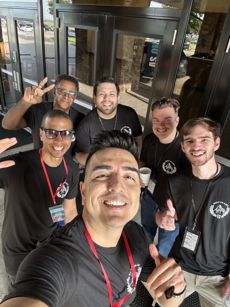 Five smiling people taking a selfie outside a building, wearing black shirts and red lanyards.