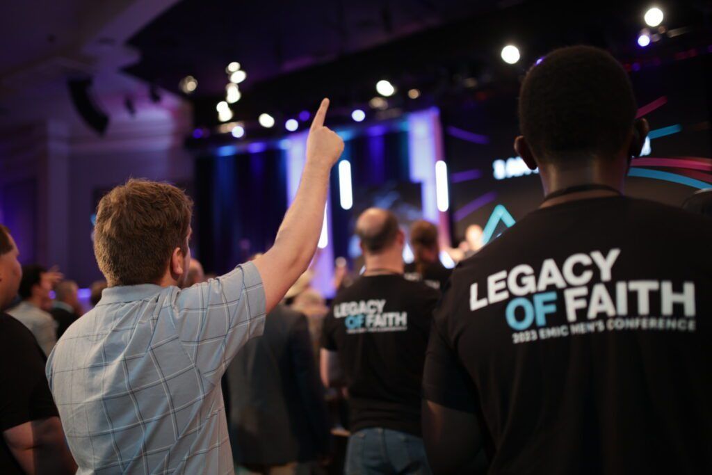 Crowd at a worship conference with raised hands under stage lights, one shirt reads “Legacy of Faith”