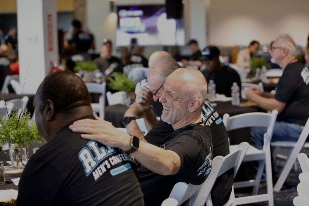 People seated in rows at an indoor event, with one man resting a hand on another’s shoulder.
