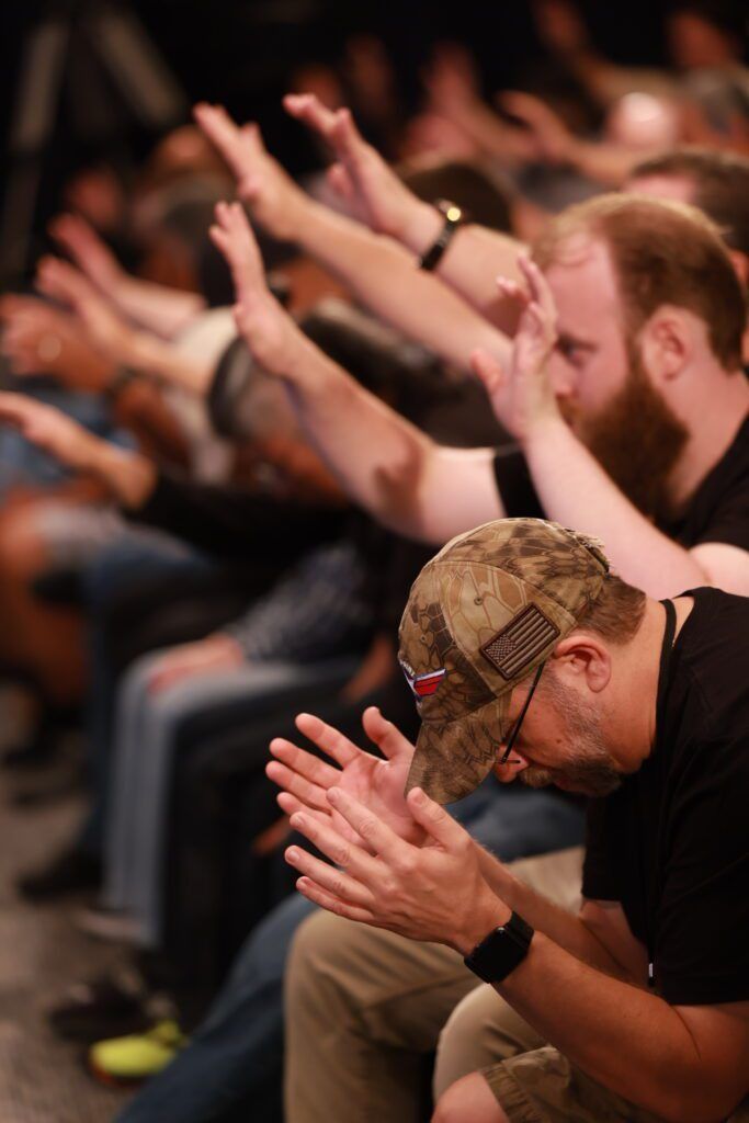 Audience members bowing heads and clapping with raised hands in a dimly lit venue