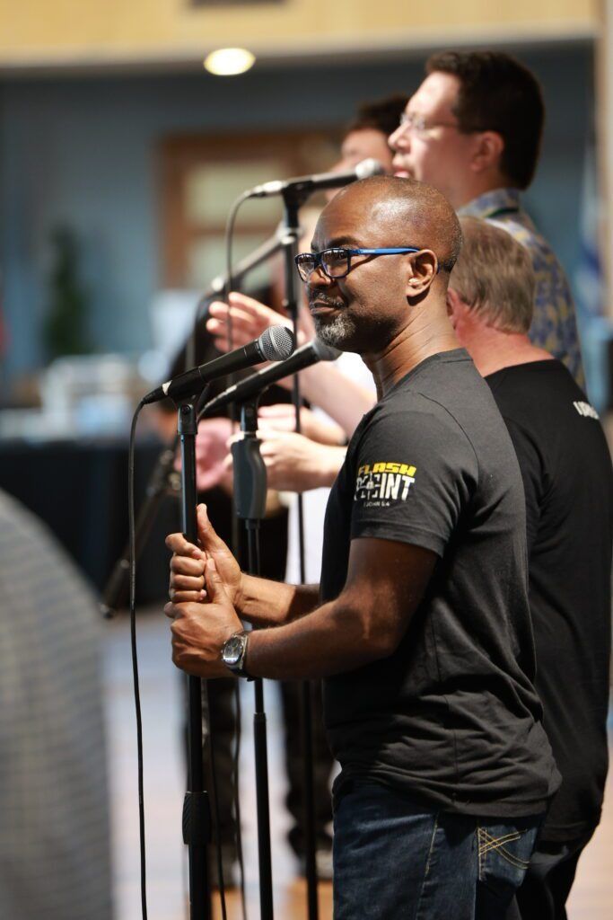 Three singers at microphones performing indoors, one in a black T-shirt in the foreground.
