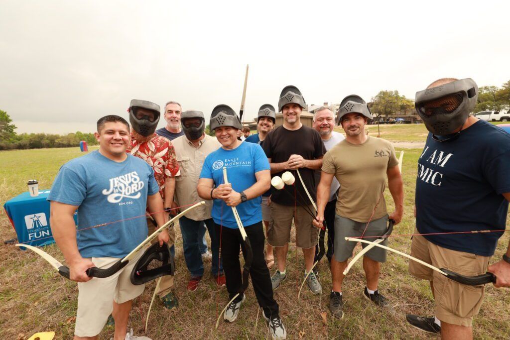 Group of people outdoors holding bows and arrows in a field, smiling at the camera.