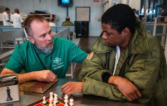 Two people leaning over a chessboard in a café, one in a green shirt and one in an olive jacket.
