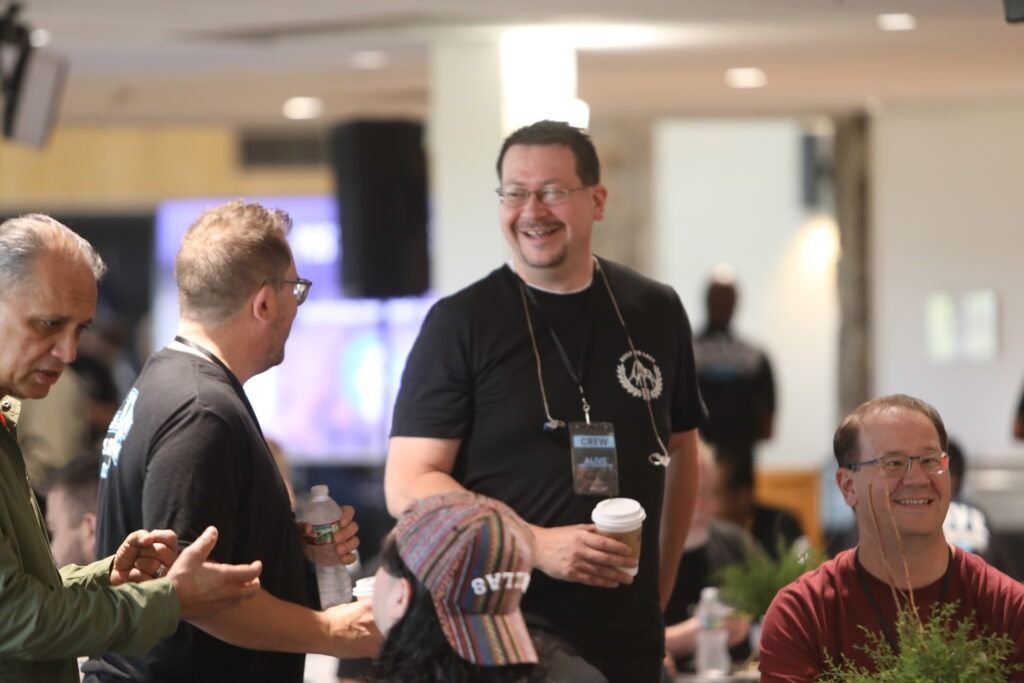 People mingling at an indoor event, with one man in black holding a coffee cup and smiling.