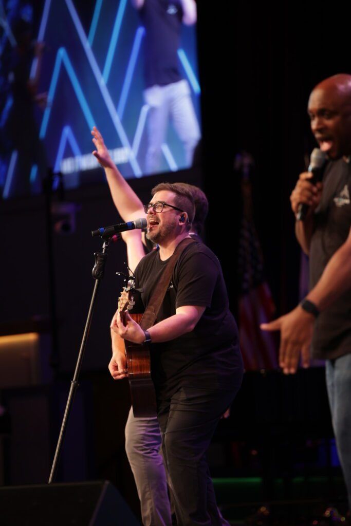 Musician singing and playing guitar onstage beside a microphone, with a large screen background