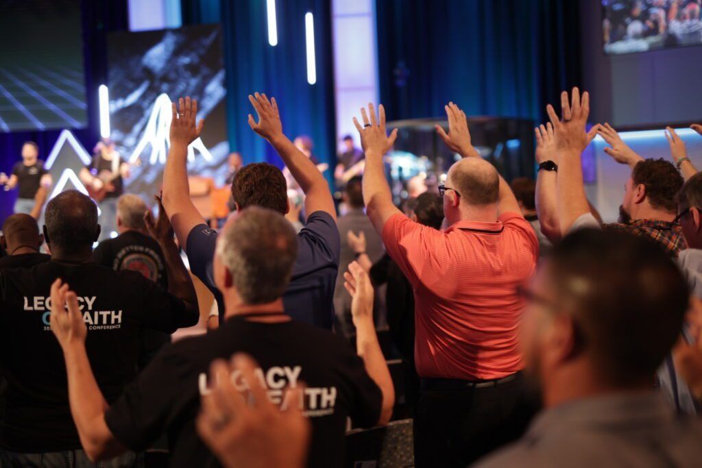 Crowd with raised hands at a church service, facing a stage with blue lighting