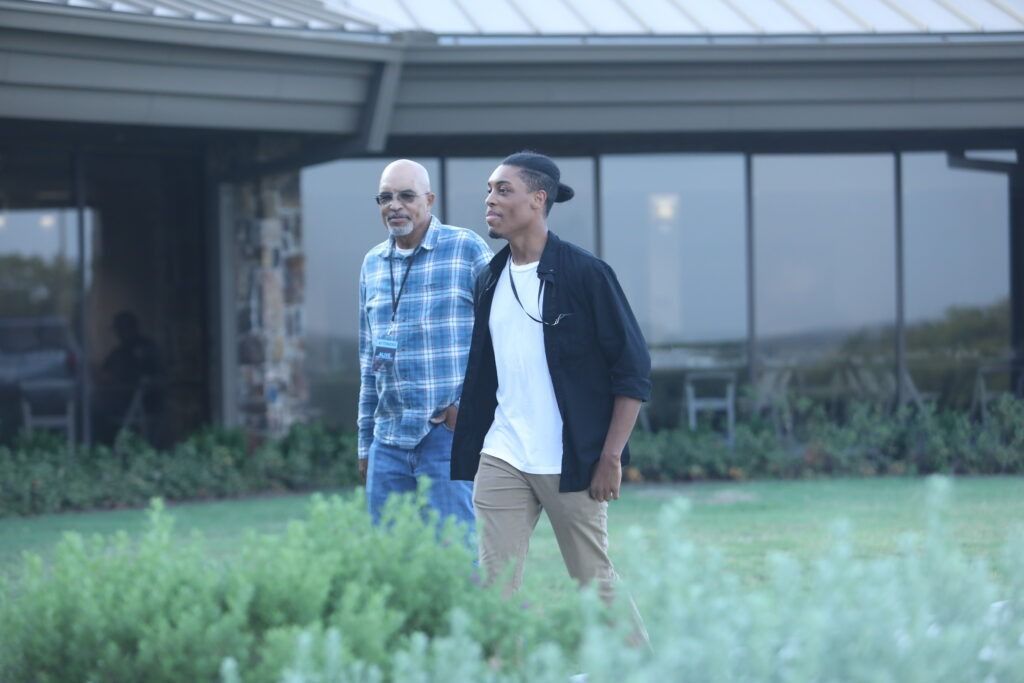 Two men walking outside a modern house, one in a blue plaid shirt and one in a dark jacket.