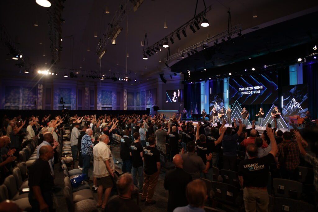 Crowd gathered in a large auditorium with stage lights and performers on a brightly lit stage