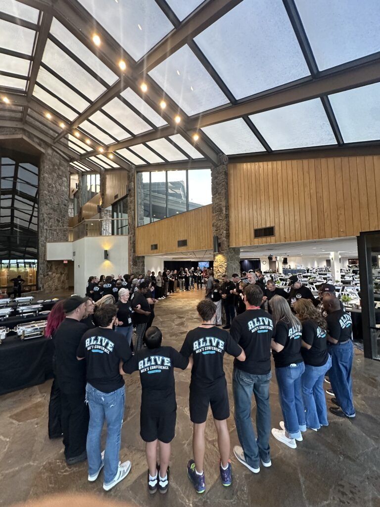 Group in black shirts stands in a bright indoor hall with a glass ceiling and wood-paneled walls.