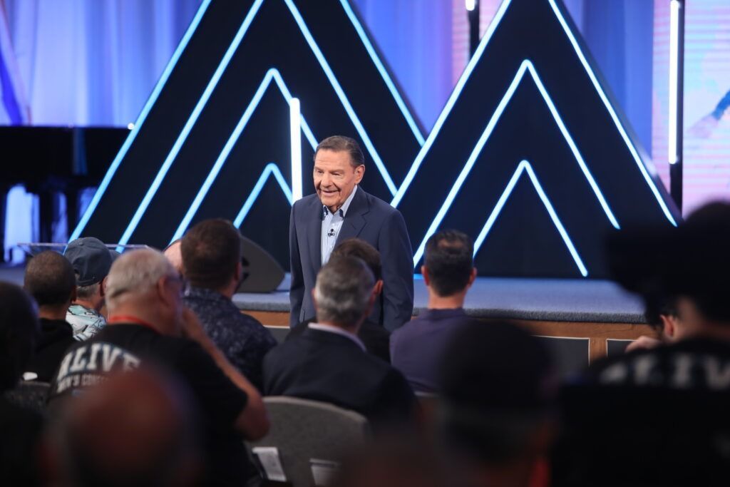 Man speaking on stage at a conference, with audience seated in front and blue geometric lights behind him