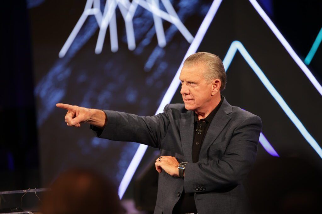 Man on stage pointing left, wearing a dark blazer against a geometric light backdrop