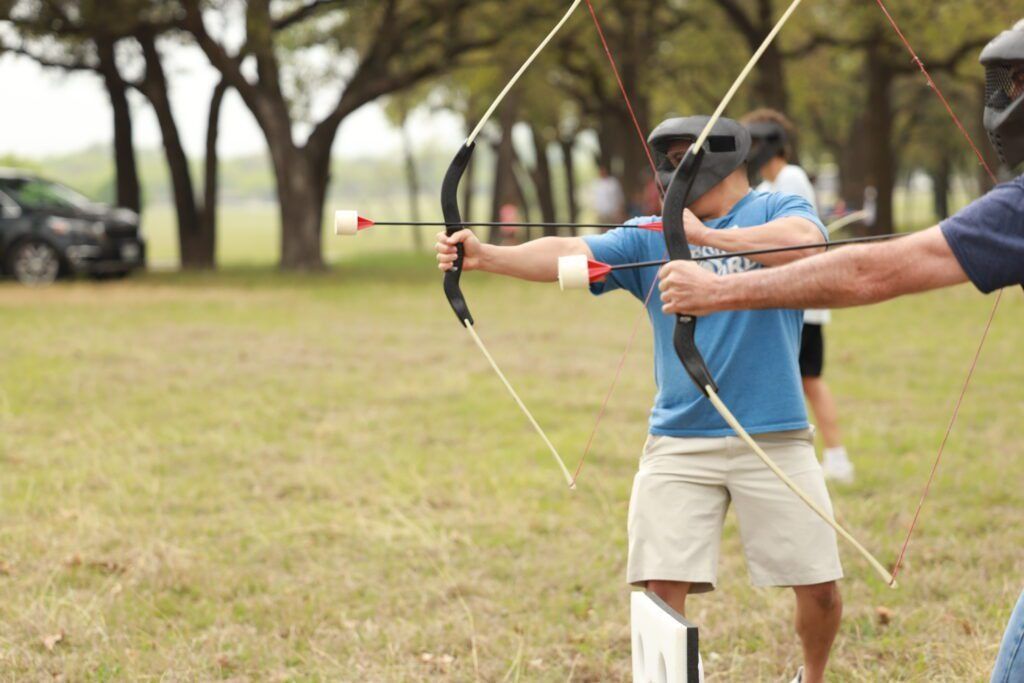 People practicing archery outdoors, aiming bows at a target in a grassy park.