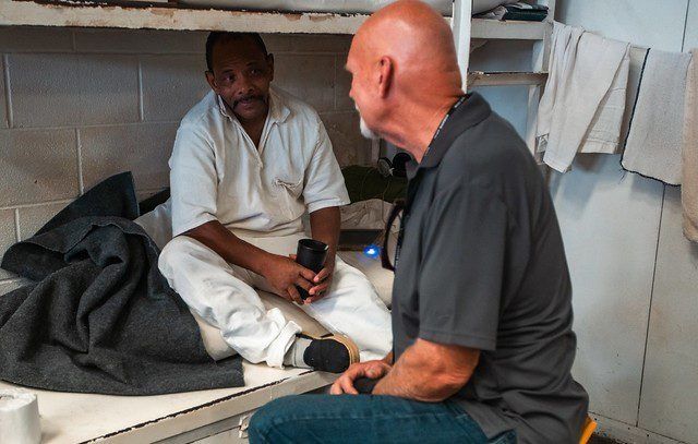 Two men seated on a bunk bed, talking in a small room with folded towels on the wall.