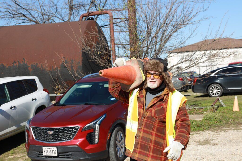 Person in an orange safety vest and fur hat stands in front of a red SUV, pointing outdoors.