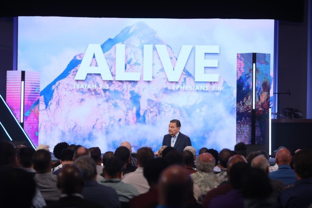 Speaker on stage before large “ALIVE” screen, addressing an audience in a dark auditorium.