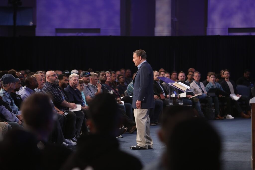 Speaker addressing a seated audience in a large conference hall under purple lighting