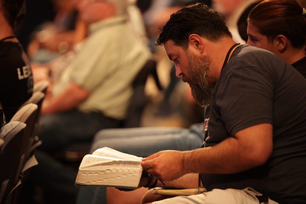 Man kneels beside a small white cake in a dim indoor gathering, head bowed in concentration.