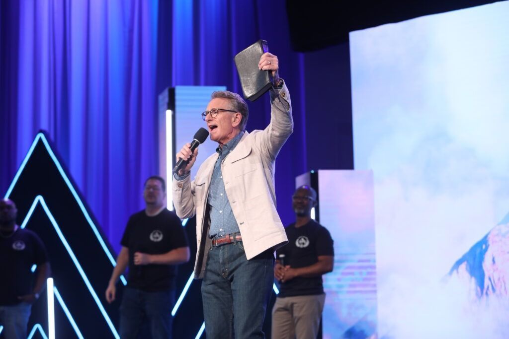 Speaker on stage holding a microphone and raising a book, with blue lights and screens behind him.