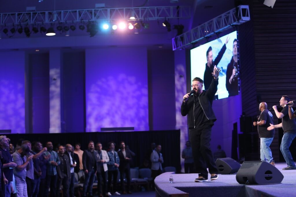 Concert stage with performer singing under purple lights, crowd watching from the side and a screen behind him