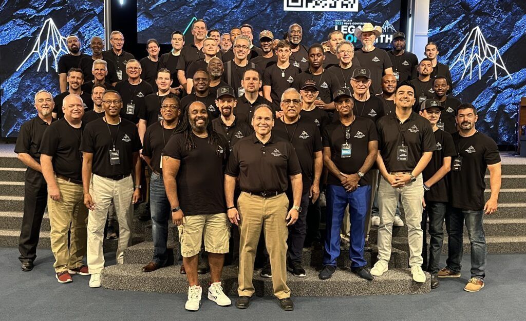 Large group in black shirts posing together on a stage in front of blue mountain-like graphics and steps