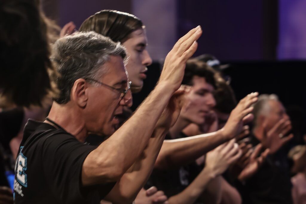Audience members seated in rows, clapping with raised hands in a dimly lit venue
