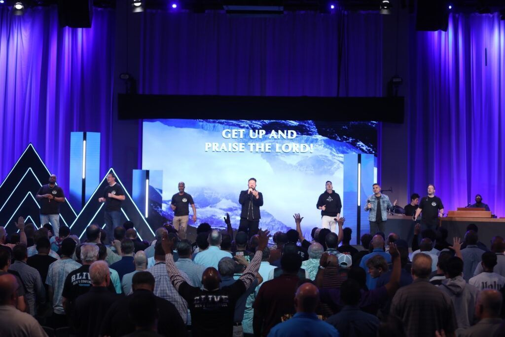 Crowd at a church worship service with stage lights and screen reading “Get Up and Raise the Energy”
