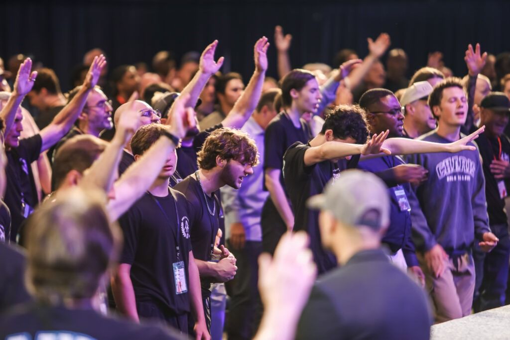 Crowd of people cheering with raised hands at a lively indoor event, lit in purple lighting