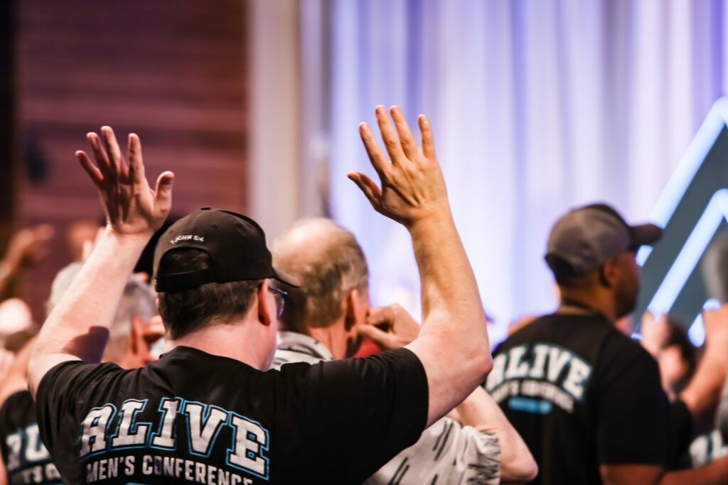Audience members raising hands at a conference, wearing black “ALIVE” shirts