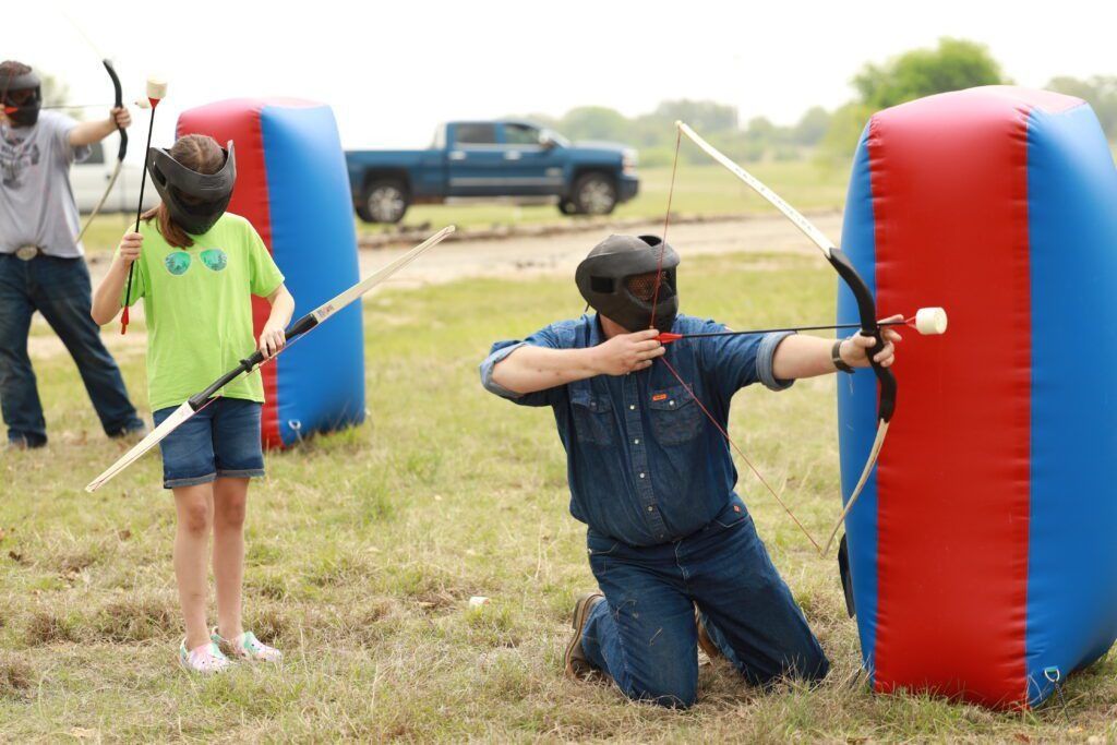 People aiming bows at targets during an outdoor archery game, with blue and red inflatable barriers.