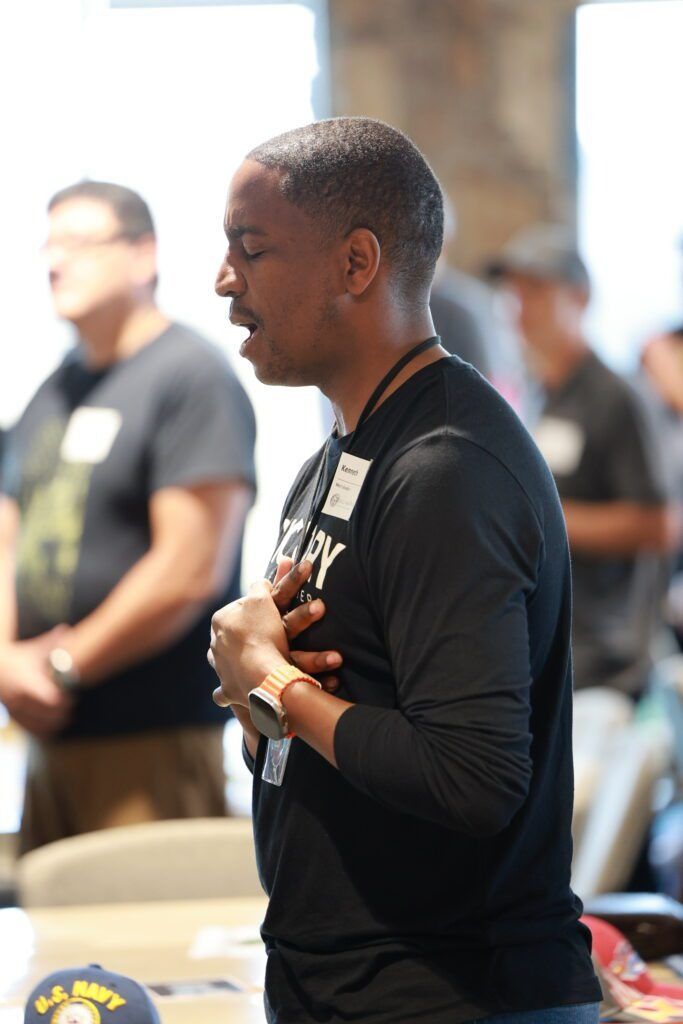 Person in a black shirt standing indoors at an event, holding their chest and looking to the side.