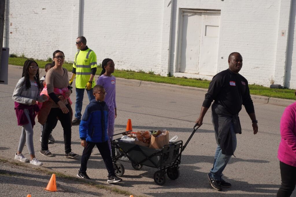 People walking with a cart and wheelbarrow on a road near a white building, with cones and a worker in a safety vest.