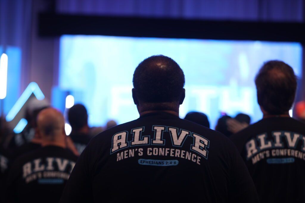 Audience wearing ALIVE Men’s Conference shirts facing a blue-lit stage