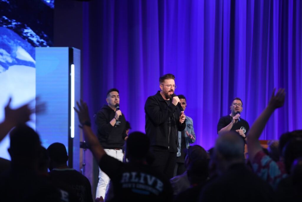 Three singers perform onstage under purple lights as a crowd raises their hands.