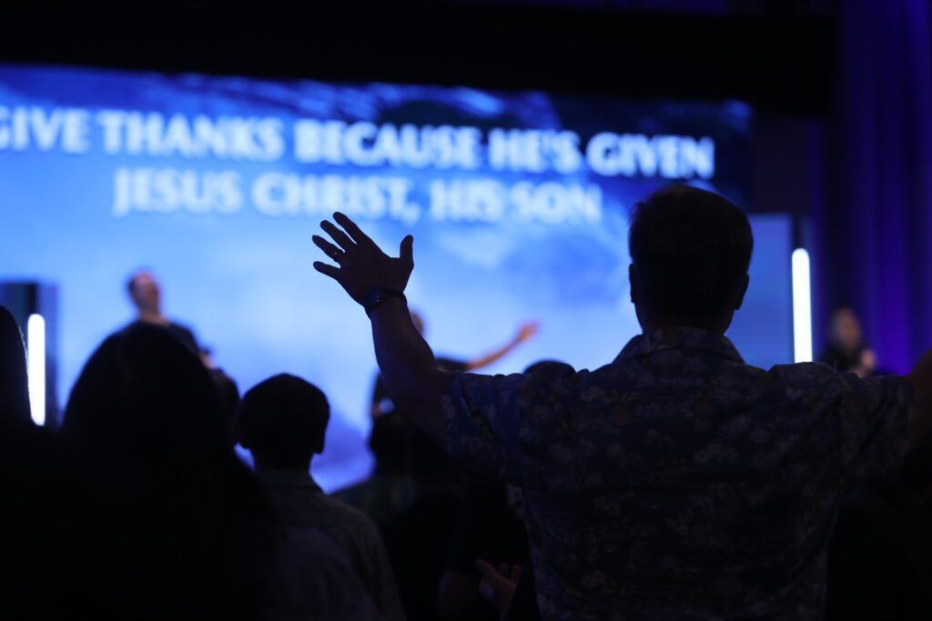 Silhouetted worshippers raising hands before a blue-lit stage with large Christian lyrics on screen