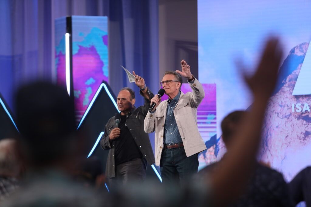 Two presenters onstage with microphones, one raising a hand, in a blue-lit event setting