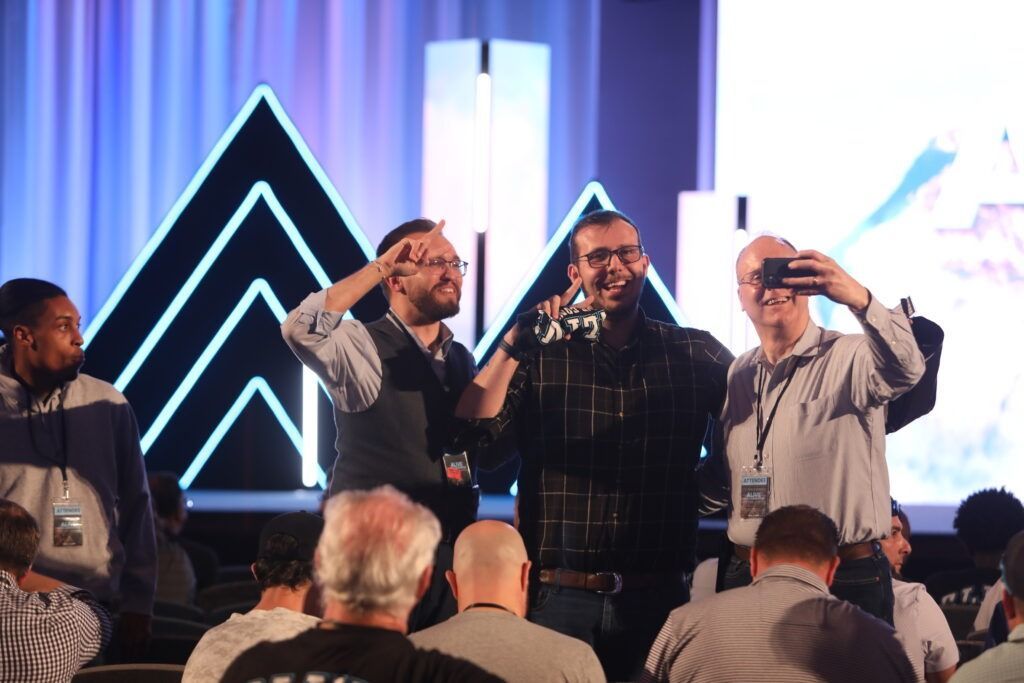 Four people onstage singing into microphones before a seated audience, with glowing neon triangle lights behind them