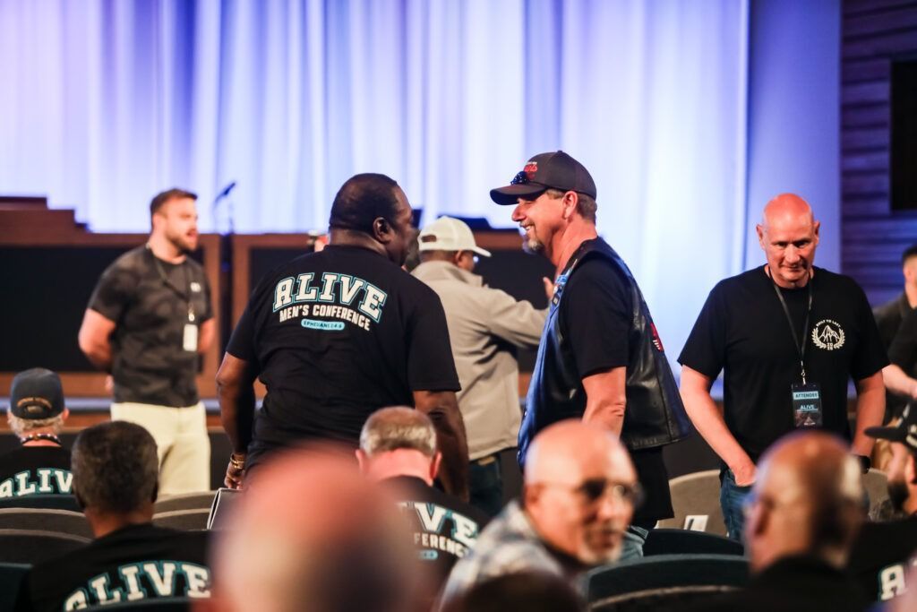 People in black shirts chatting at an indoor event with a white curtain backdrop