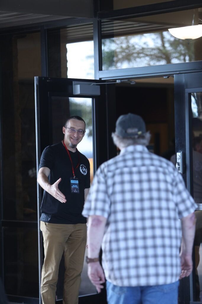 Man in a black shirt greeting a man in a plaid shirt at an open doorway.