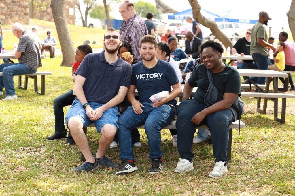 Three friends sitting on a picnic table bench outdoors in a park, smiling at the camera.