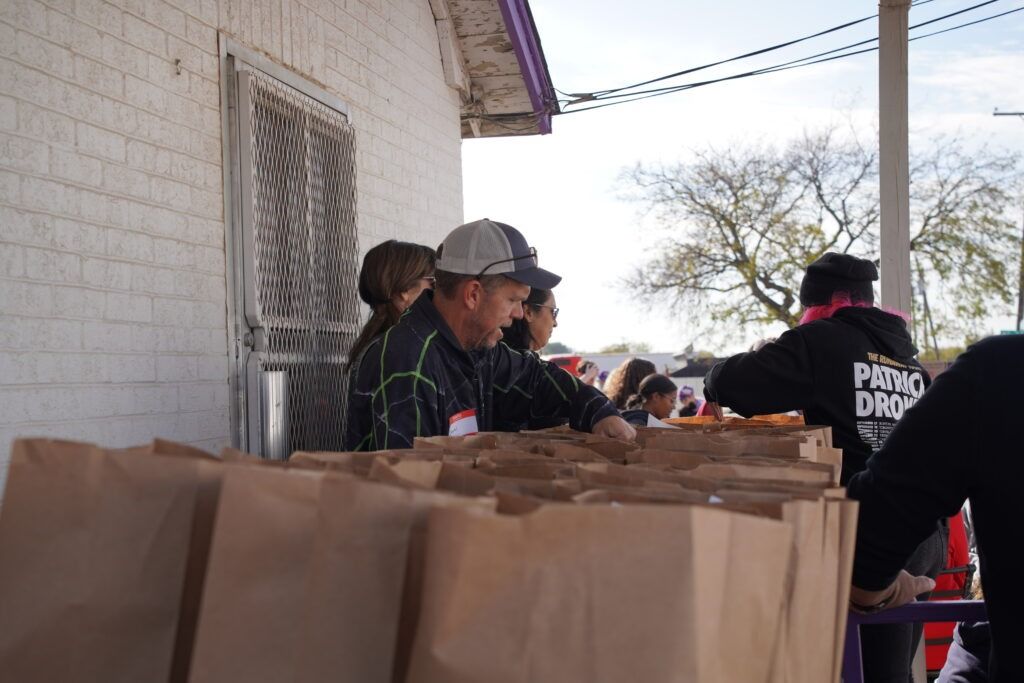 Volunteers handing out brown paper bags from a table beside a white brick building outdoors