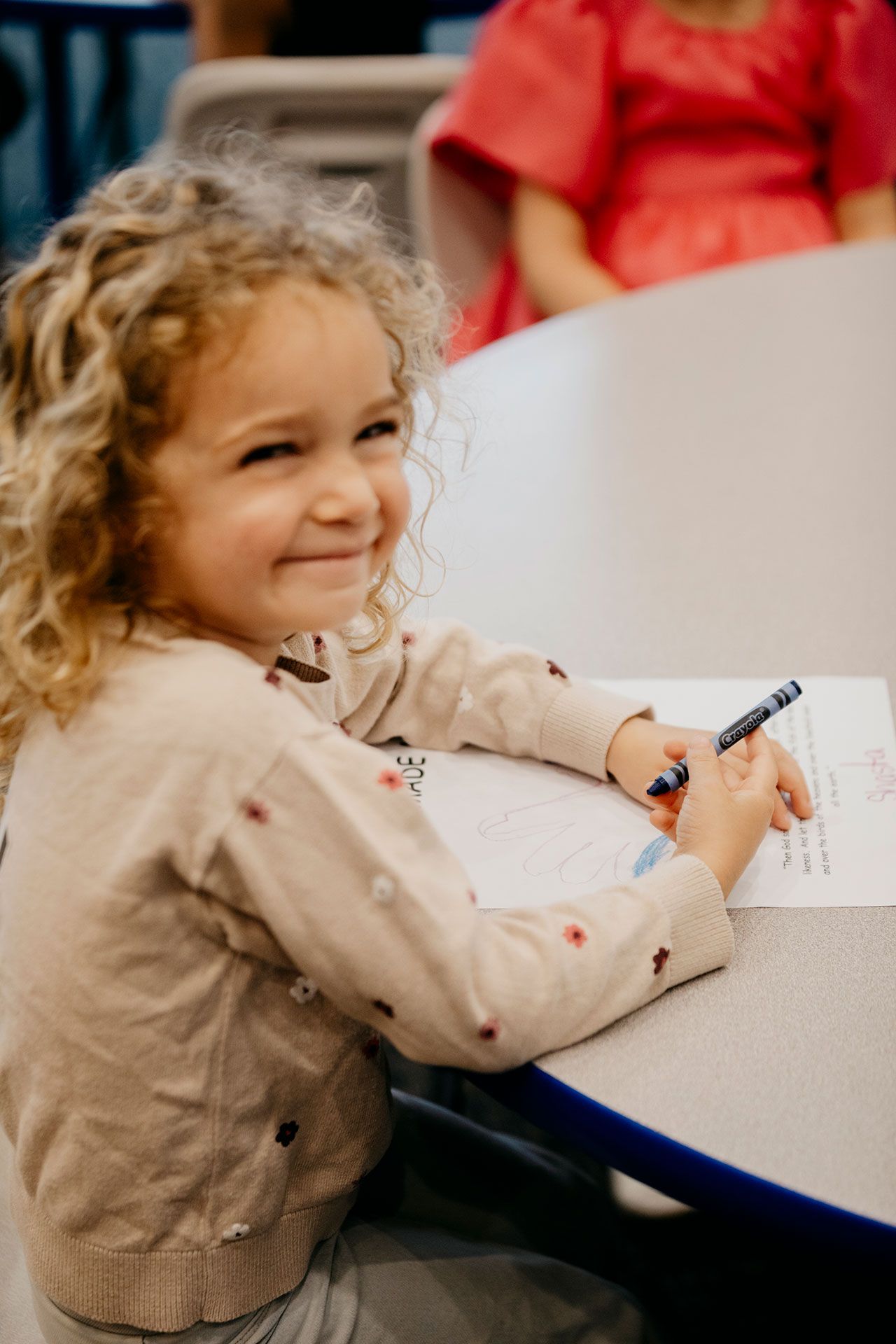 Child smiling and drawing with crayons at a white table in a classroom setting