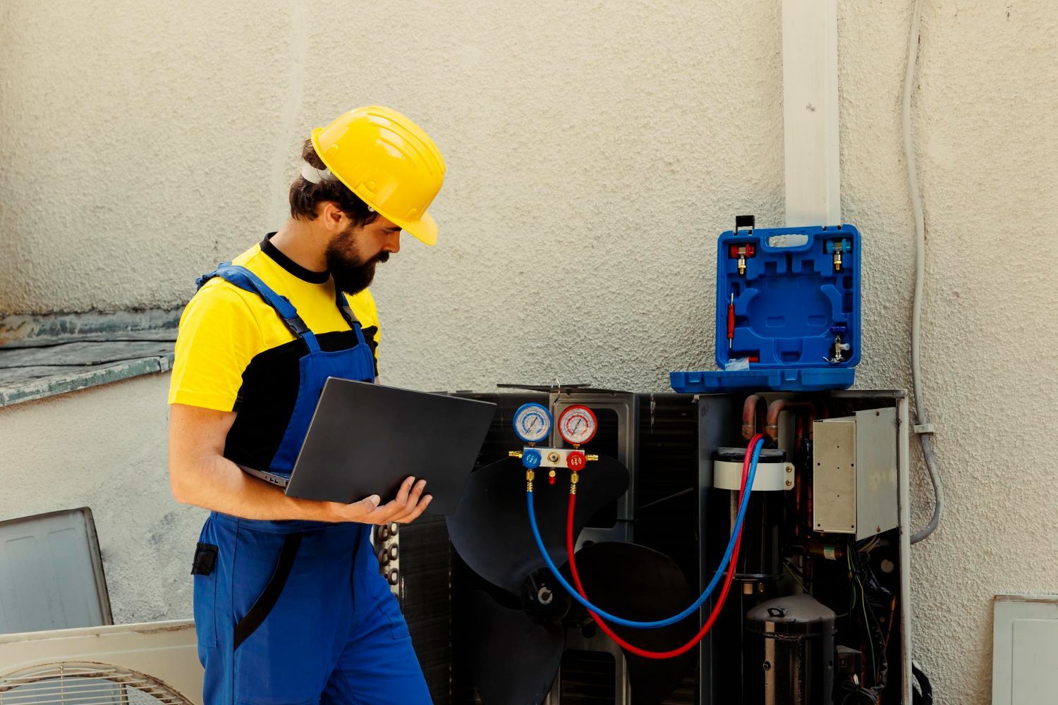 A man wearing a hard hat and overalls is working on an air conditioner.