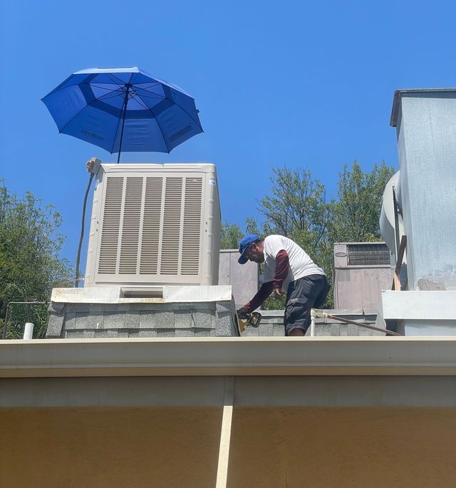 A worker on a rooftop near an air conditioning unit, using a tool under a blue umbrella on a sunny day.