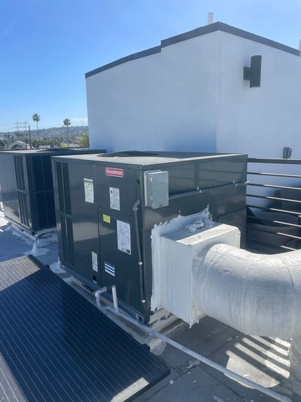Two rooftop HVAC units on a flat roof under a bright blue sky, near a white building.